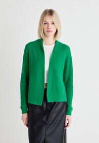 Gilet en tricot côtelé vert avec un devant ouvert et des manches longues, associé à un haut blanc et une jupe en cuir noir. Texture lisse, design décontracté.
