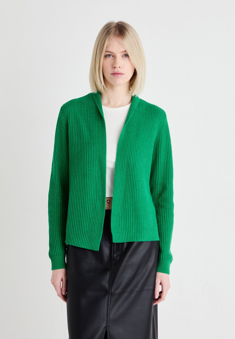 Gilet en tricot côtelé vert avec un devant ouvert et des manches longues, associé à un haut blanc et une jupe en cuir noir. Texture lisse, design décontracté.