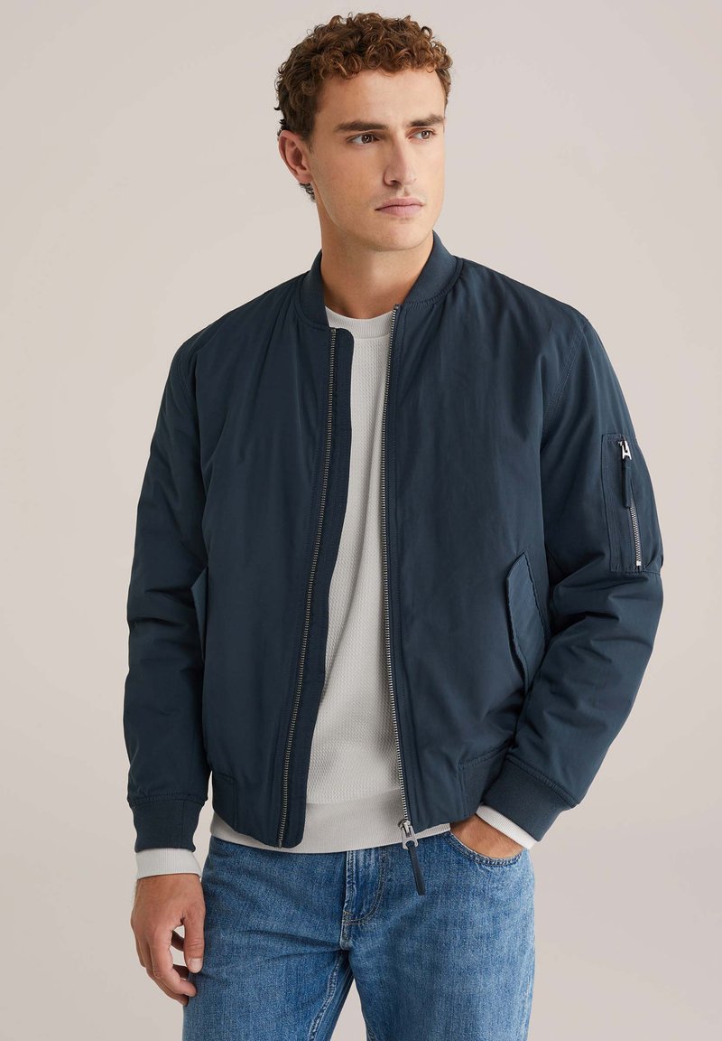Giacca bomber blu navy con zip frontale, tasche laterali e polsini a costine. Materiale liscio e leggero con una vestibilità moderna e aderente.