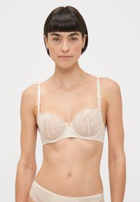 Soutien-gorge beige à armatures avec des détails en dentelle florale, des straps ajustables et des accents décoratifs sur l'épaule, présentant un design en tulle transparent.