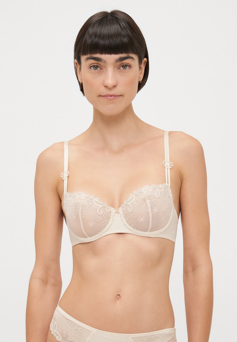 Beige underwire bra with floral lace detailing, adjustable straps, and decorative accents at the shoulder, featuring a sheer mesh design.