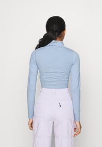 Helles blaues Langarm-Turtleneck-Oberteil kombiniert mit lavendelfarbenen Cargo-Hosen, die Taschen und ein Logodetail auf der Rückseite haben. Glatte Textur.