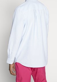 Chemise à manches longues bleu clair avec plis au dos, poignets boutonnés et ourlet arrondi, en tissu doux, associée à un pantalon rose.