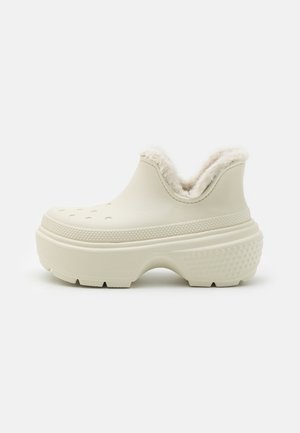 Sabots beige clair, hauteur cheville, avec une doublure en peluche, un bout rond et des trous de ventilation, présentant une semelle texturée et une construction robuste.