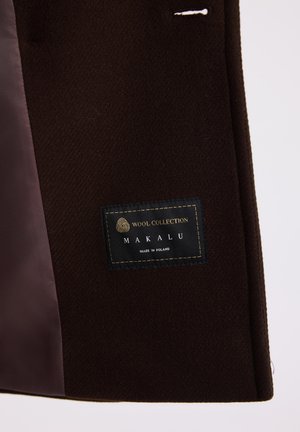 Wełniana marynarka w ciemnym brązie o teksturowanej powierzchni. Posiada przyszytą czarną metkę ze złotym napisem, informującym "Wool Collection Makalu, wyprodukowane w Polsce."