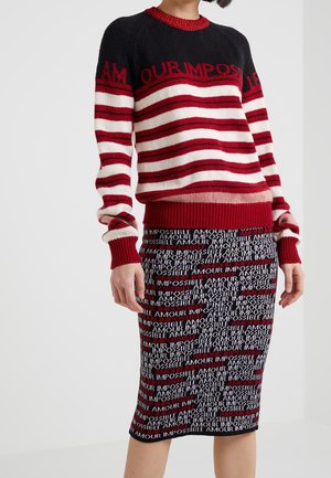 Femme portant un pull rayé rouge, blanc et noir et une jupe noire avec le texte répété « AMOUR IMPOSSIBLE » en rouge et blanc.