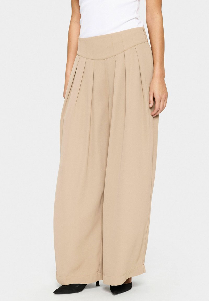 Pantaloni beige a gamba larga con pieghe, una texture di tessuto liscia, vita alta e tasche laterali. Abbinati a tacchi neri a punta.