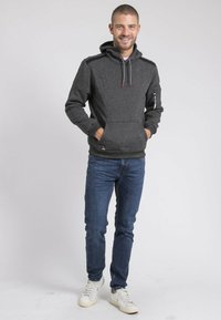 Sweat-shirt gris à capuche avec un tissu texturé, poche avant et accents rouges. Porté avec un jean bleu et des baskets blanches.