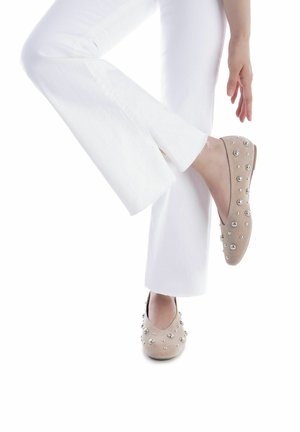 Zapatos planos de ante beige con punta redondeada, adornados con tachuelas circulares plateadas, combinados con pantalones blancos acampanados. Textura suave, diseño minimalista.