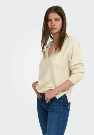 Ung kvinde med langt brunt hår iført en cremefarvet V-hals sweater og blå jeans, stående med hænderne i lommerne mod en ensfarvet baggrund.