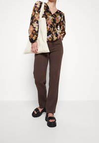 Blumenmuster Langarmbluse mit geknoteter Vorderseite, kombiniert mit braunen, hochgeschnittenen Hosen und klobigen schwarzen Sandalen. Weiße Tragetasche in der Hand gehalten.