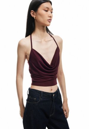 Top sin mangas burdeos con un profundo escote en V y diseño drapeado, combinado con unos jeans de tiro alto azul oscuro, que realzan las texturas y líneas suaves.