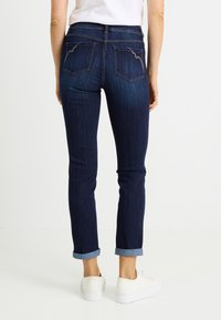 Vaqueros de mezclilla azul oscuro con pierna cónica y puños remangados. Presentan dos bolsillos traseros con un distintivo detalle de costura.