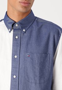 Camicia a bottoni blu e bianca con texture denim, taschino sul petto e piccolo logo nei colori rosso, bianco e blu. Colletto classico.