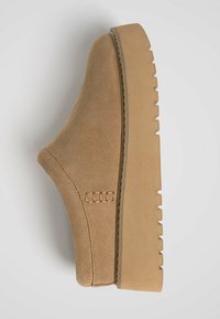 Beige suede slip-on shoe with a thick, ridged sole, featuring a woven accent on the side and a rounded toe design.