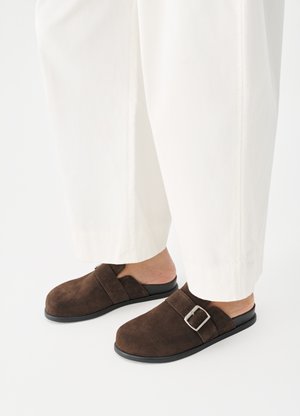 Személy fehér nadrágot és sötétbarna, ezüst csattal díszített velúr félcipőt visel, egy sima fehér felületen áll.