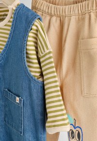 Denim overal s náprsním vreckom, pruhovaná dlhá košeľa v zelenej a krémovej farbe a béžové nohavice so grafickým motívom jablka a textúrovanou látkou.