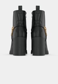 Crne kožne čizme do gležnjeva s debelim potplatom, s dekorativnim remenom i zlatnim detaljima. Glatka tekstura s minimalnim dizajnom.