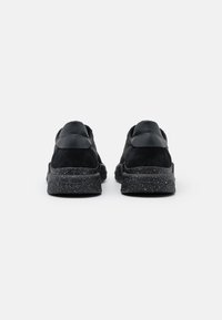 Une paire de baskets noires avec des semelles noires tachetées, vues de l'arrière sur un fond blanc uni.