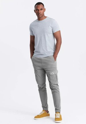 Camiseta gris de cuello redondo, pantalones cargo grises con bolsillos laterales y un parche logo. Zapatillas amarillas de corte bajo con suelas blancas. Atuendo simple y casual.
