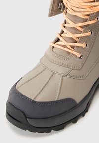 Beige vanntette støvler med grå gummitupp, teksturert læroverdel, kontrasterende oransje snørebånd og metalløyler. Forsterket design for holdbarhet.