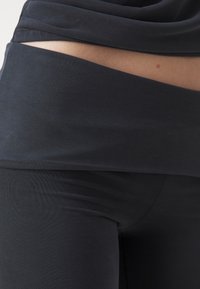 A fekete leggings rétegezett derékrész kialakítással, sima textúrával és testhezálló szabással rendelkezik, hangsúlyozva a letisztult, minimalista megjelenést.