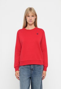 Felpa rossa con collo e polsini a coste, caratterizzata da un piccolo logo a cuore nero sul petto. Indossata con jeans blu.