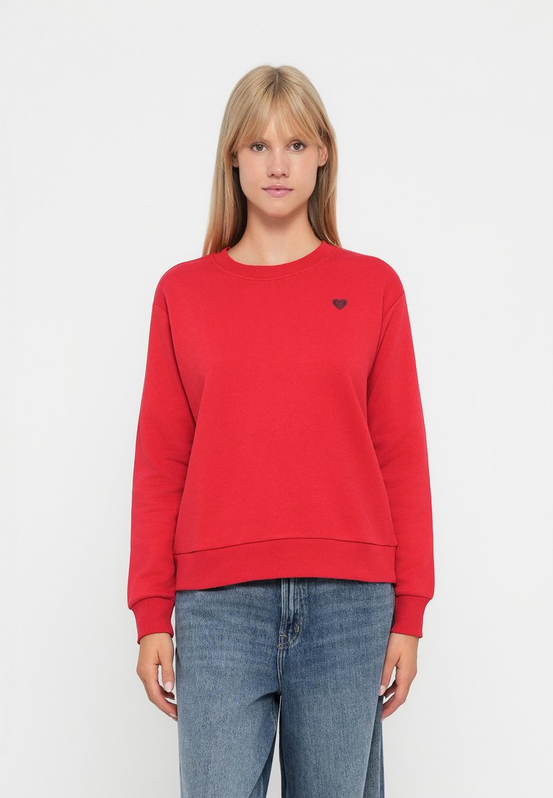 Felpa rossa con collo e polsini a coste, caratterizzata da un piccolo logo a cuore nero sul petto. Indossata con jeans blu.