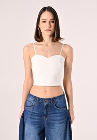 Top corto bianco con scollo a cuore e spalline sottili, abbinato a jeans di denim vita alta con spacchi laterali e vestibilità rilassata.
