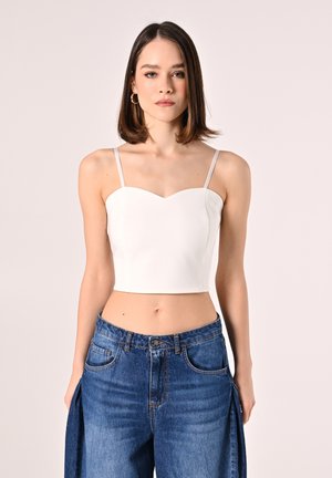 Weißes Crop-Top mit Spaghettiträgern und Herz-Ausschnitt, kombiniert mit hochgeschnittenen blauen Jeans mit seitlichen Schlitzen und Taschen.