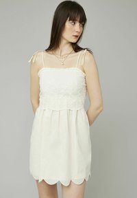 Robe blanche avec des bretelles fines réglables, broderies florales sur le corsage, ourlet festonné et coupe décontractée. Le tissu texturé semble léger.