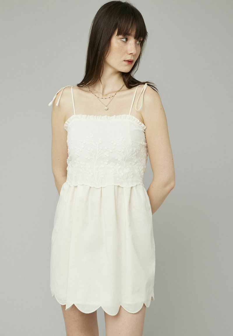 Robe blanche avec des bretelles fines réglables, broderies florales sur le corsage, ourlet festonné et coupe décontractée. Le tissu texturé semble léger.