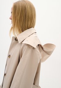 Beige trenčkot s elegantním designem, velkým límcem, mírně zdůrazněnými rameny a zapínáním na jeden knoflík.
