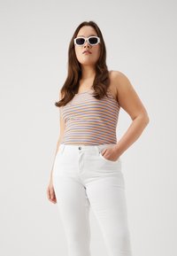 Top de tirantes a rayas en naranja y azul, diseño ajustado; combinado con jeans skinny blancos. Modelo lleva gafas de sol blancas con un fondo neutro.