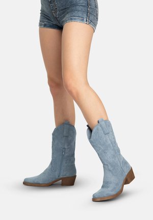 Gambe con stivali da cowboy in camoscio blu a metà polpaccio con tacchi bassi e pantaloncini di denim blu su uno sfondo semplice.