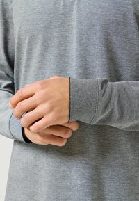 Szara koszula z długim rękawem o teksturowanym wzorze. Rękawy mają kontrastowy wzór. Tkanina wydaje się być miękka i lekka. Ręce są delikatnie złożone.