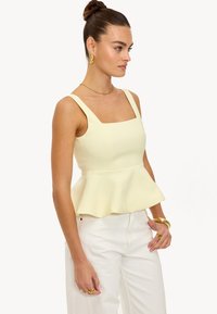 Top peplum jaune avec encolure carrée et larges bretelles, associé à un pantalon blanc taille haute. Les accessoires comprennent des boucles d'oreilles et des bagues en or.