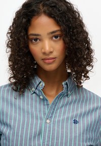 Chemise à rayures bleues et blanches avec un col, présentant des rayures d'accent vertes et un petit logo brodé. Texture de tissu douce.