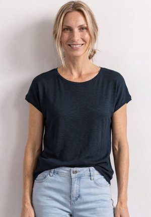 Uśmiechnięta kobieta z blond włosami związanymi z tyłu, ubrana w czarną koszulkę z krótkim rękawem i jasnoniebieskie dżinsy, stojąca na tle gładkiej białej ściany.