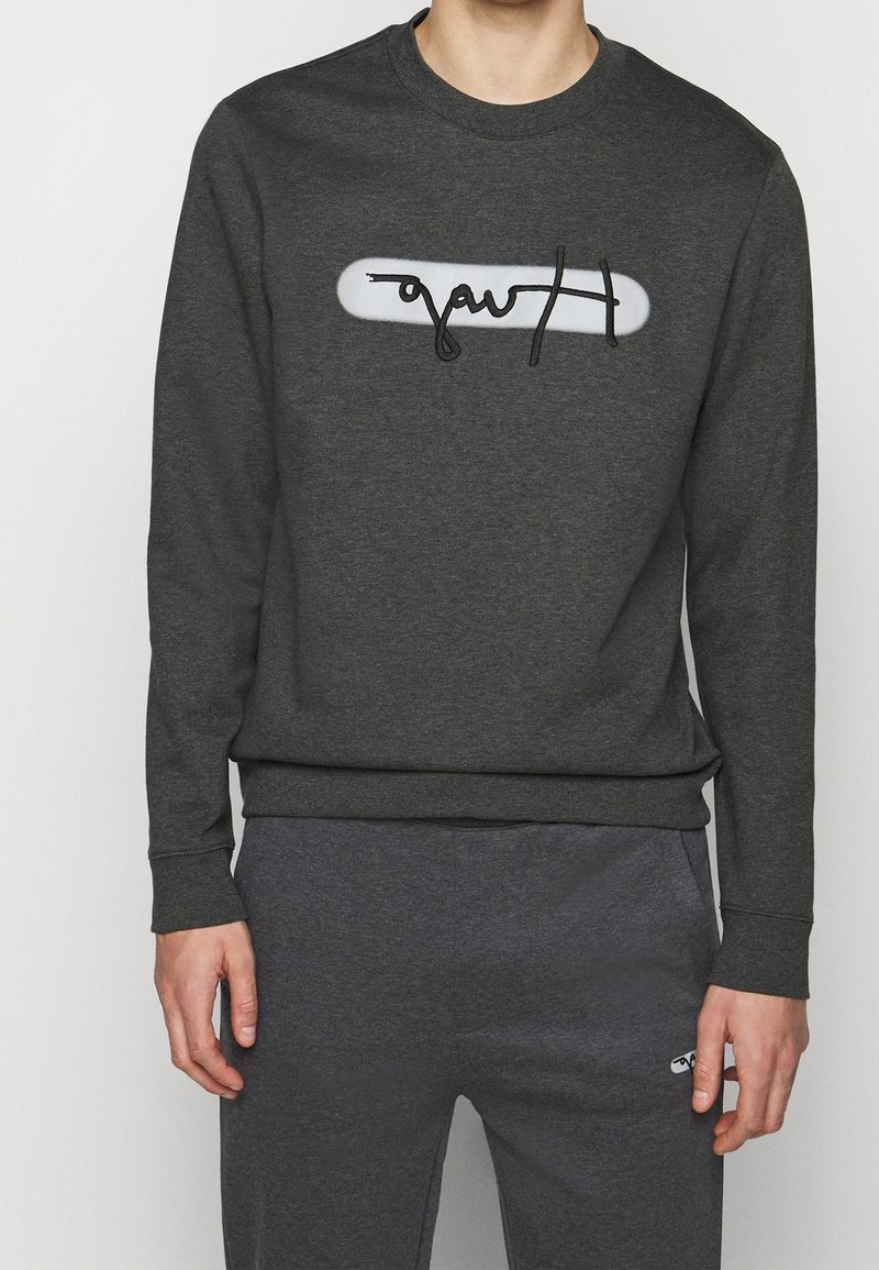 Sudadera gris oscuro con cuello redondo, que presenta un panel rectangular blanco y un diseño de logo negro en el centro. Textura de tela suave.