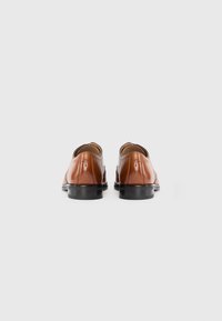 Zapatos de cuero marrón con un diseño elegante, que presentan un tacón bajo y suela de goma negra. En la parte trasera se aprecian costuras sutiles y un detalle de marca.