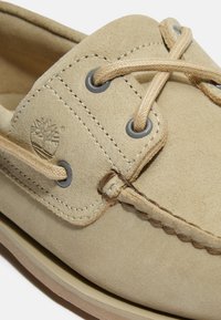 Chaussure bateau en daim beige avec lacets beiges, œillets métalliques, coutures détaillées et logo en relief de l’arbre Timberland sur le côté.