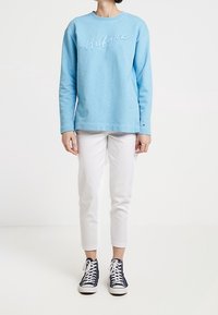 Sudadera azul clara con "Hilfiger" bordado en blanco, combinada con pantalones ajustados blancos y zapatillas marinas. Textura suave y diseño de cuello redondo.