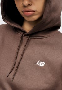 Καφέ φούτερ με κορδόνι, απαλό ύφασμα και λευκό λογότυπο "New Balance". Διαθέτει μπροστινή τσέπη καγκουρό και ελαστικά τελειώματα στα μανίκια.