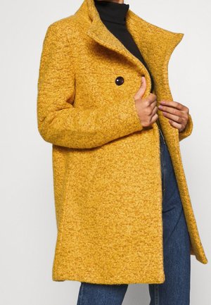 Woman wearing a textured mustard yellow wool coat over a black turtleneck and blue jeans, holding the coat open with one hand.