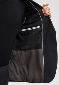 Zwart colbert met grijs gestructureerde stof, voorzien van een gladde binnenvoering, een ritssluiting aan de zak en contrasterende witte accenten op de randen.