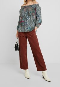 Ilmava, olkapäiltä avoin blouse, jossa on monivärinen paisley- ja ympyräkuvio, yhdistettynä ruosteenvärisiin leveälahkeisiin corduroy-housuihin ja valkoisiin nilkkureihin.