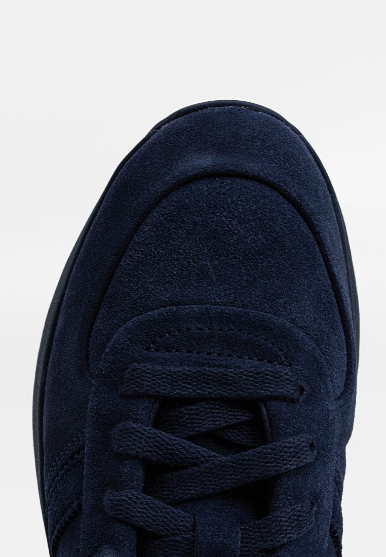 Gros plan sur l'avant d'une sneaker en suède bleu marine avec des lacets assortis et des détails de couture sur un fond blanc propre.