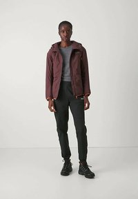 Γυναίκα φορώντας μια σκούρα μπορντό τζάκετ με κουκούλα πάνω από γκρι μπλούζα, μαύρες φόρμες και μαύρα αθλητικά παπούτσια, που στέκεται μπροστά σε απλό ανοιχτό φόντο.