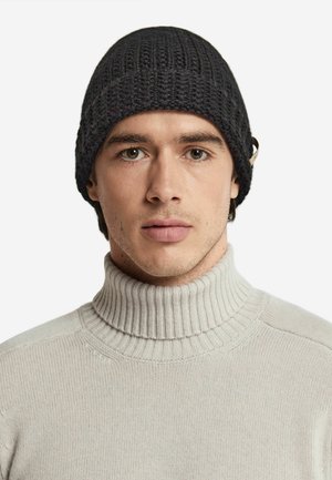 Knit black beanie with a textured pattern, featuring a small accent on the side. Worn with a light gray turtleneck sweater.